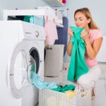 Young Smiling Woman Smelling Clothes After Washing In Washing Machine At Utility Room